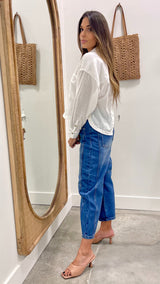 Woman in a white blouse and blue jeans standing in front of a mirror with wicker bags on the wall.