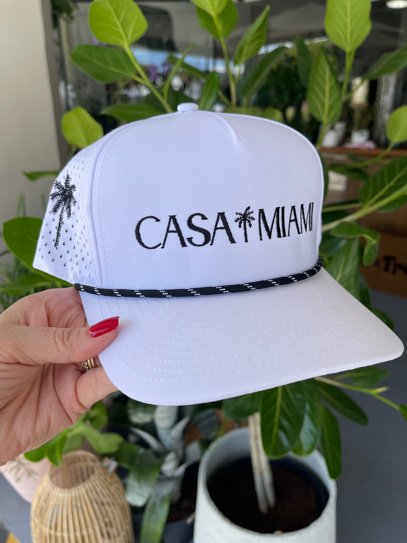 White hat with 'CASA MIAM' text held by a hand against a blurred indoor background