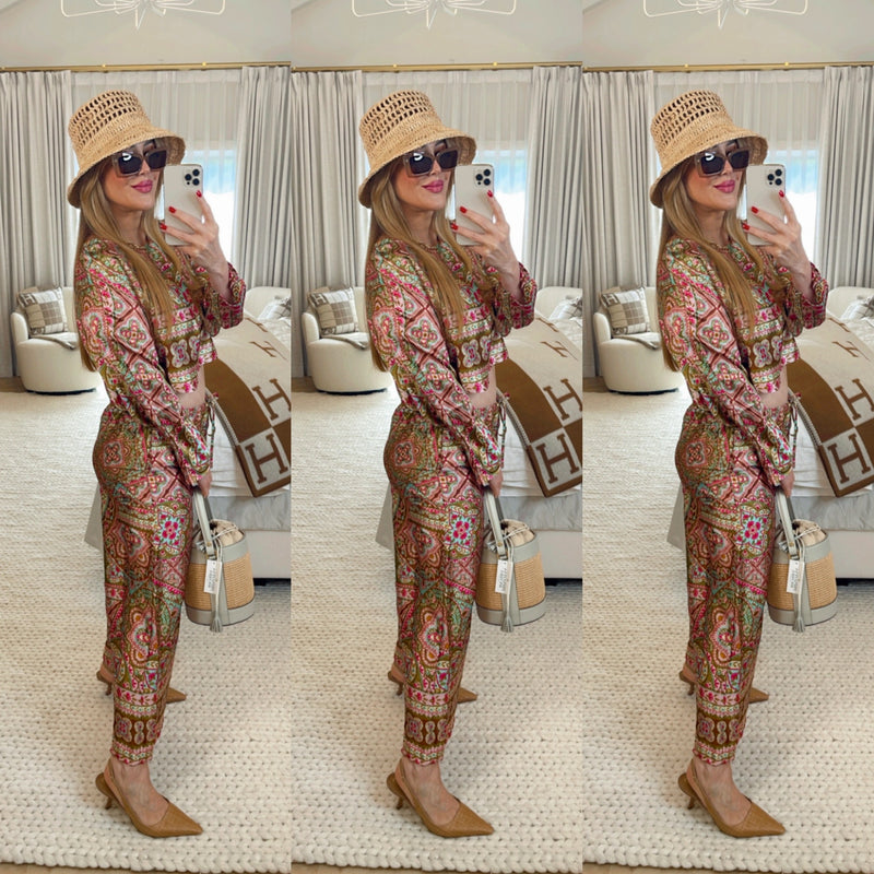 Woman in a patterned outfit with a hat and sunglasses, holding a bag, in a room with a couch and rug.