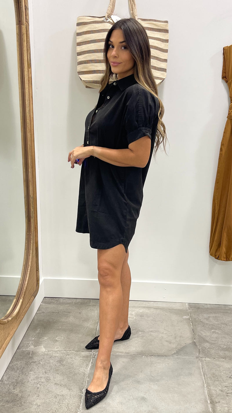 Woman wearing a black dress standing in front of a mirror with a striped bag on her shoulder.