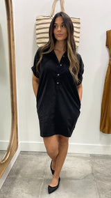 Woman wearing a black shirt dress standing in a room with a mirror and striped bag in the background.