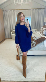 Woman in a blue dress and brown knee-high boots taking a mirror selfie in a stylish room.