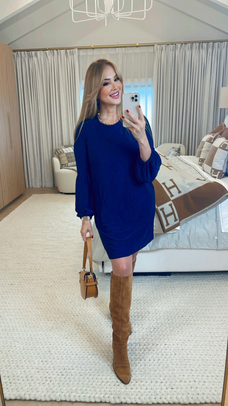 Woman in a blue dress and brown knee-high boots taking a mirror selfie in a stylish room.