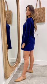Woman in a blue dress standing in front of a mirror with a wicker bag in the background.