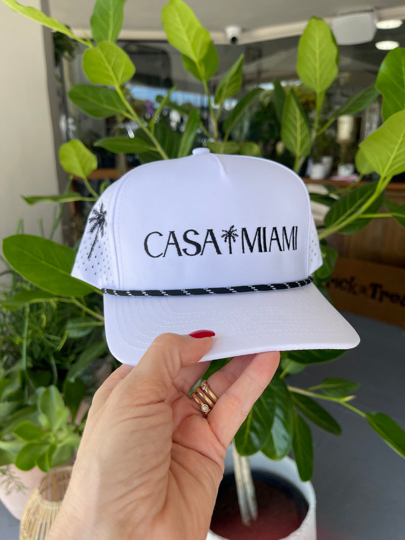 Hand holding a white mask with 'CASA MUMI' branding in front of green foliage.