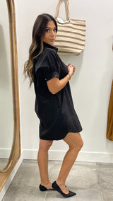 Woman wearing a black dress standing in front of a mirror with a striped bag on a white wall.
