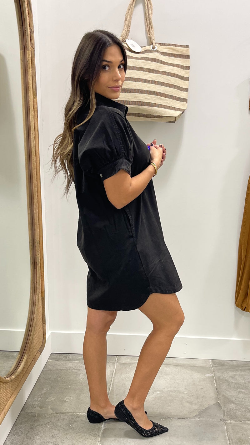 Woman wearing a black dress standing in front of a mirror with a striped bag on a white wall.