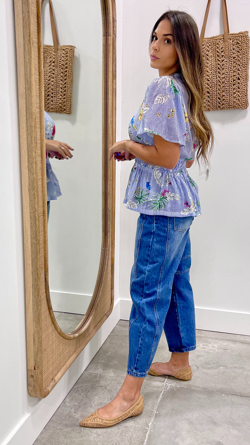 Woman in a floral blouse and blue jeans standing in front of a mirror.