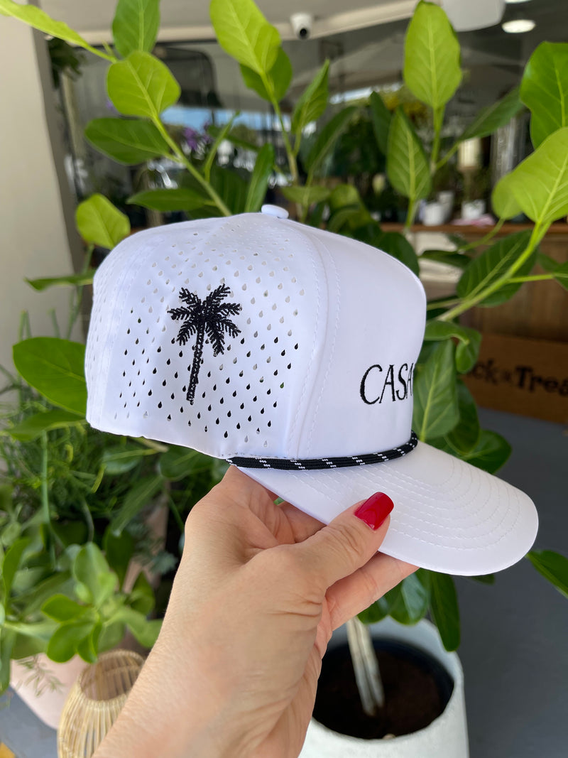 White sneakers with black palm tree design and 'CAS' branding held by a hand in front of green plants.