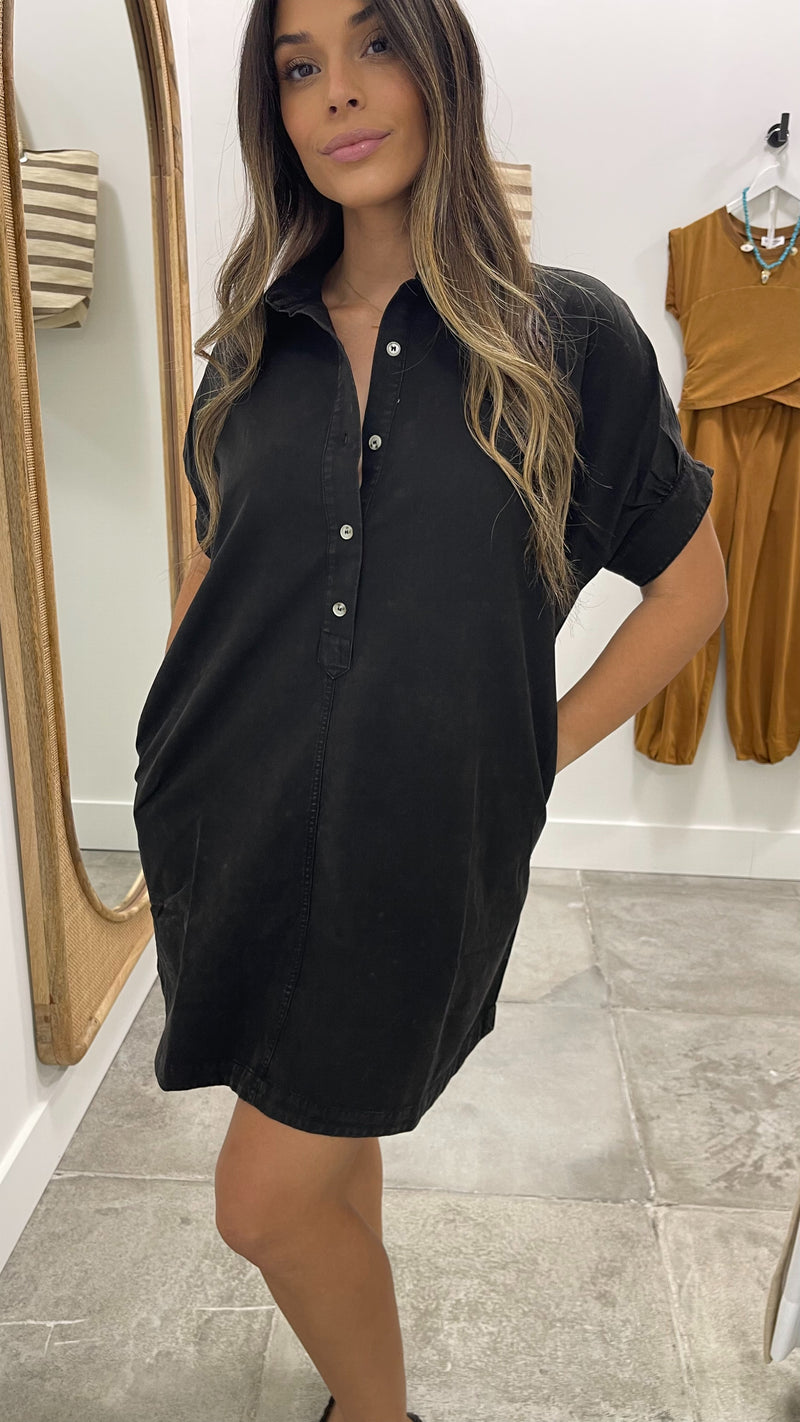Woman wearing a black dress standing in a room with a mirror and another outfit in the background.
