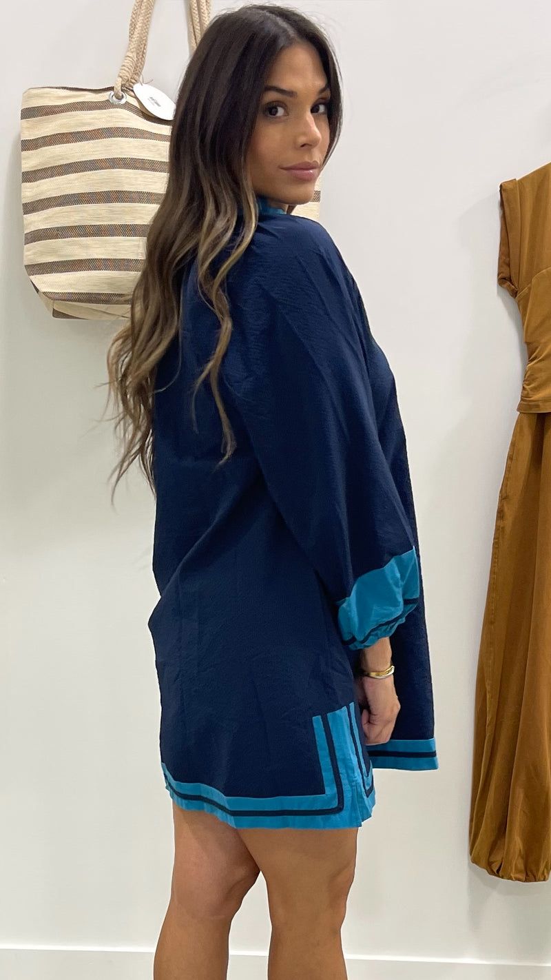 Woman wearing a navy dress with blue trim standing against a white wall.